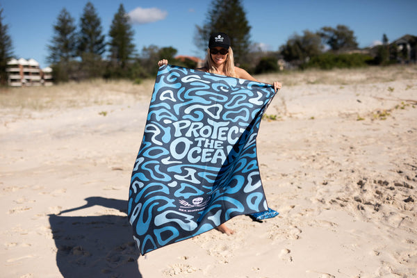 Blue and white abstract patterned recycled beach towel with "Protect The Ocean" text, displayed on sand.