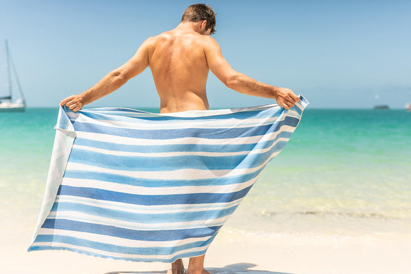 Blue and white striped recycled beach towel held open by man on sandy beach with ocean backdrop