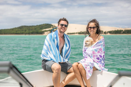 Image of a couple wrapped in Nakie's couple combo recycled sand-free beach towel while sitting on a boat, enjoying a cozy and sustainable maritime adventure.