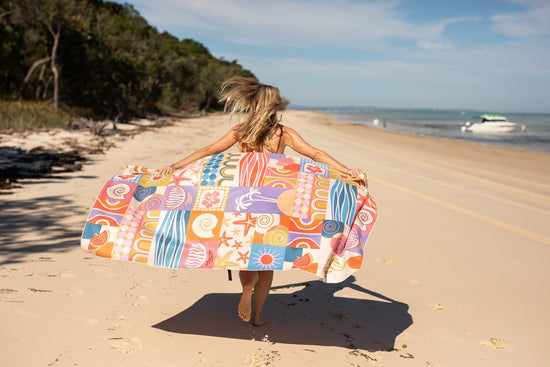 Woman walking along the beach holding up a Nakie sand-free beach towel with ocean waves in the background.