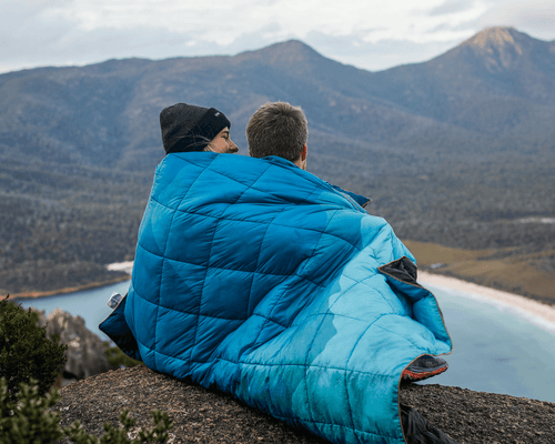 Two people wrapped in a blue puffy blanket sitting on a mountain edge