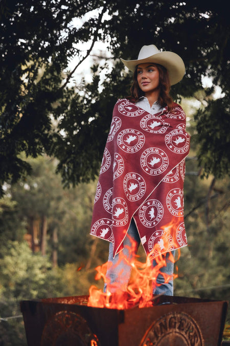 Burgundy and cream circular logo patterned beach towel draped over woman wearing cream cowboy hat by campfire
