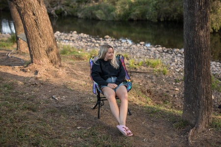 Woman sitting in the Kasey Rainbow Camping Chair by a river