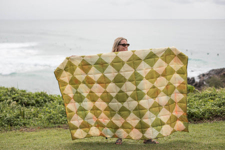 Retro Shores sustainable down puffy blanket in olive, cream, and peach diamond pattern, displayed outdoors by ocean.