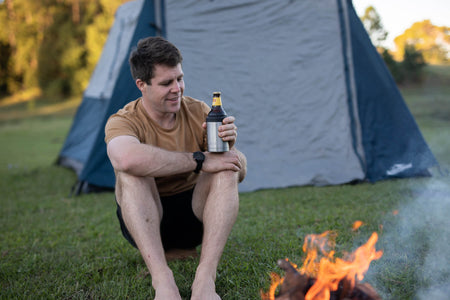 Stainless steel can cooler holding a beer bottle at camping site with tent and campfire
