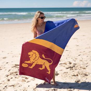 Brisbane Lions beach towel with navy, gold and burgundy color-blocking and gold lion emblem, held by woman on sandy beach