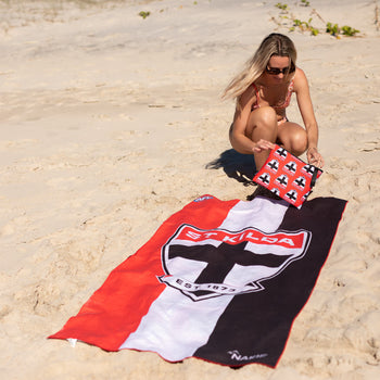 St Kilda Saints AFL beach towel in red, white and black with emblem, displayed on sand with woman holding matching pillow.