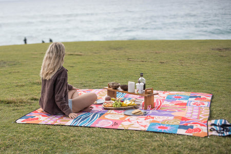 Colorful patterned picnic blanket with retro geometric design, spread on grass by ocean during seaside picnic setup.