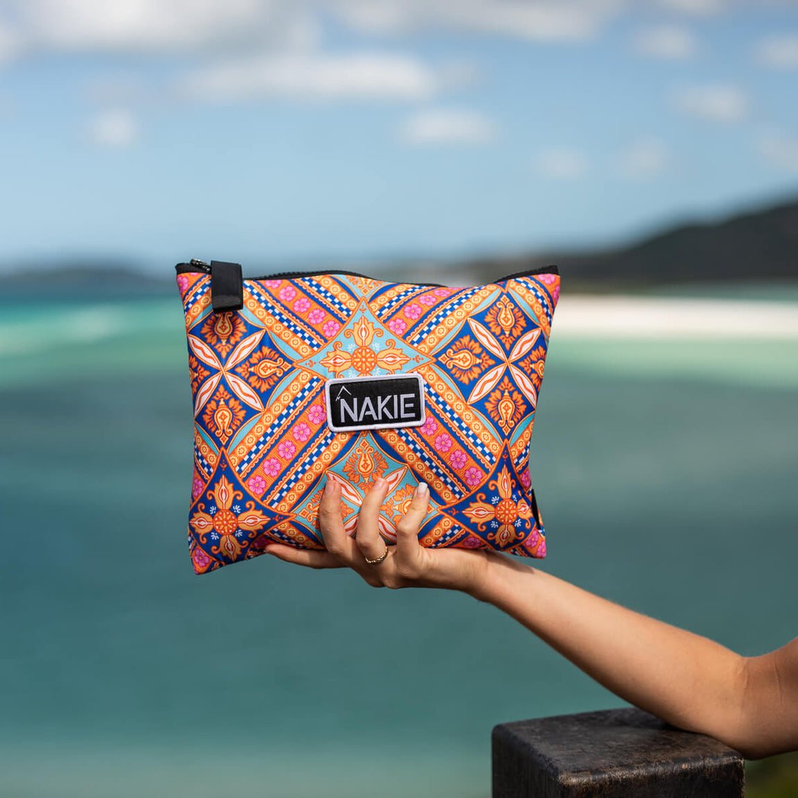 Colorful geometric patterned beach towel in orange, blue, and pink held up against a tropical ocean backdrop