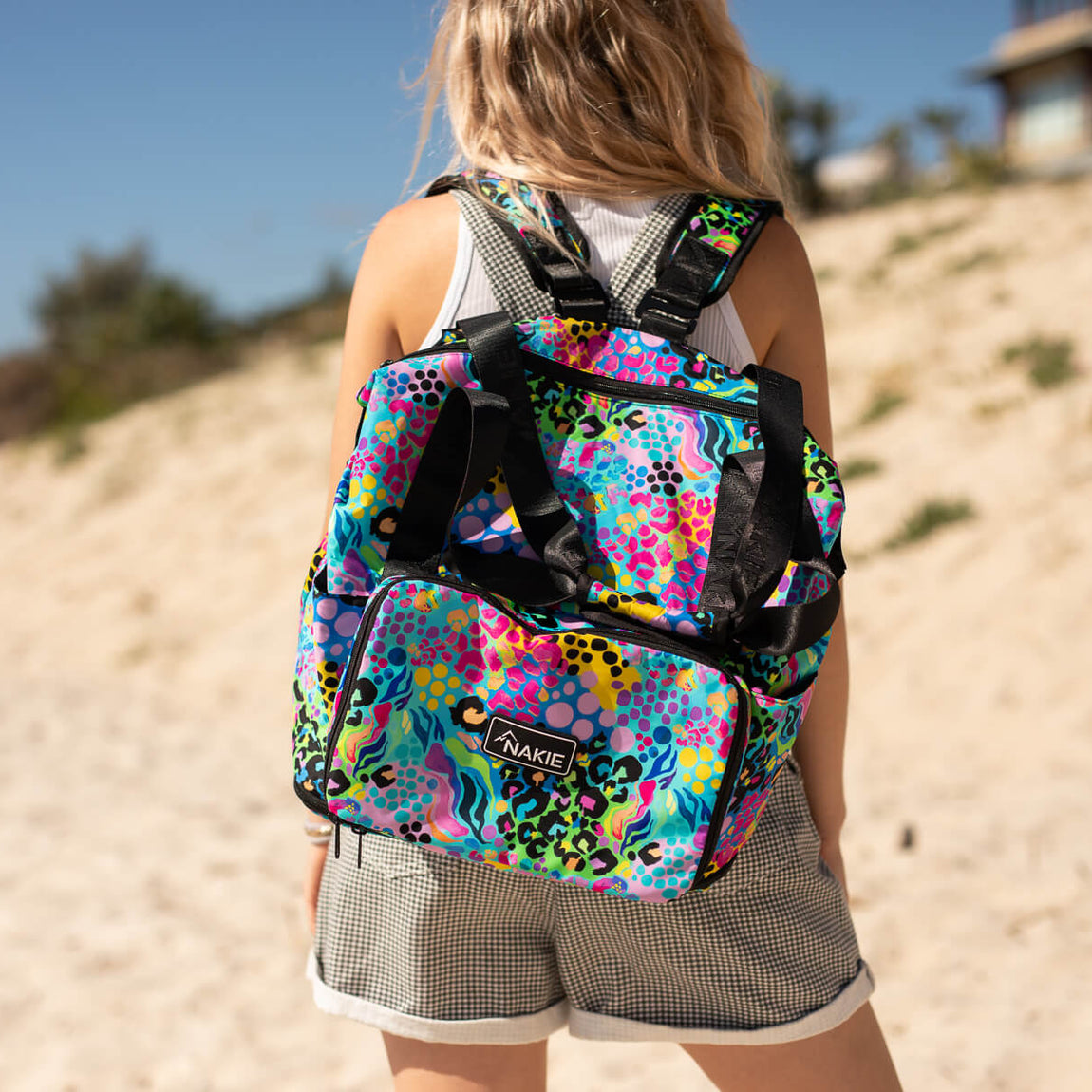 Multicolored neon geometric patterned tote backpack with black straps worn on shoulders at beach.