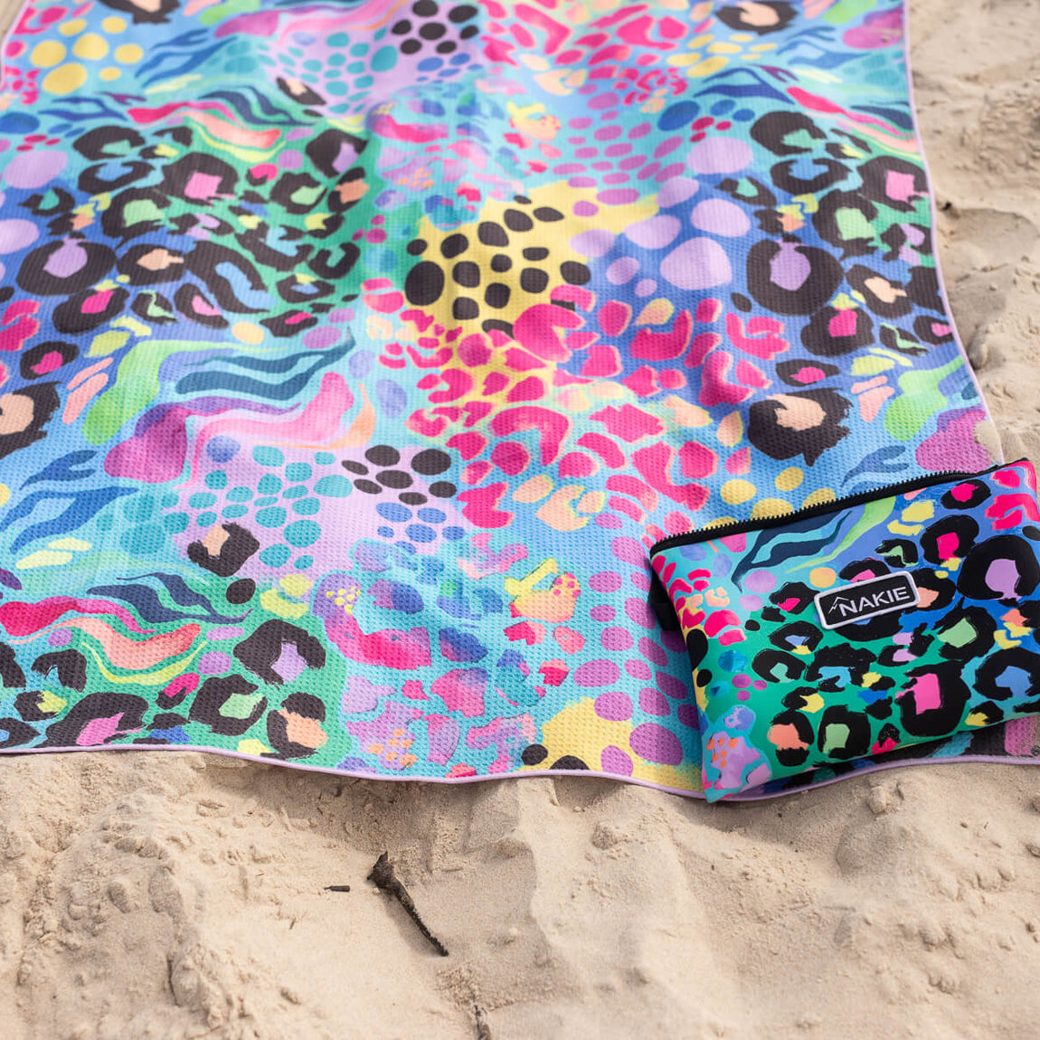 Multicoloured recycled sand-free beach towel with abstract leopard and wave pattern, displayed on sand with matching pouch.