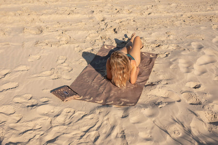Brown recycled beach towel with embroidered detail, laid on sand with person resting on it