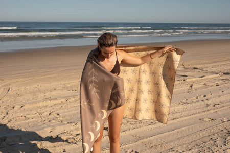 Beige recycled beach towel with moon and star print, displayed on beach by woman wrapping around shoulders