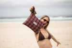 Burgundy recycled beach towel with circular logo pattern, held up at beach with black handle detail visible. preview #11
