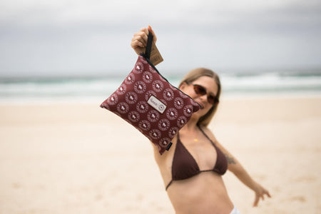 Burgundy recycled beach towel with circular logo pattern, held up at beach with black handle detail visible.