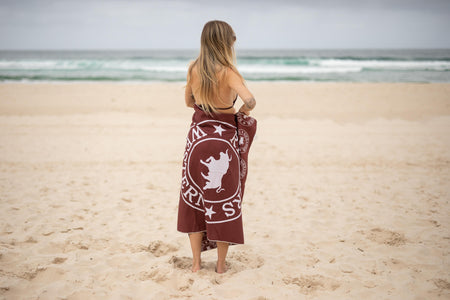 Burgundy beach towel with white graphic logo and star design, worn around woman's waist on sandy beach