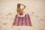 Burgundy recycled beach towel with white logo print, laid flat on sand with woman sitting on it from above. preview #7