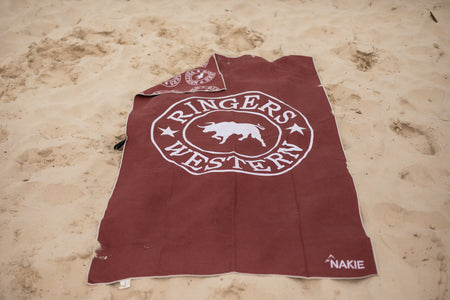 Burgundy beach towel with white Ringers Western logo and pig graphic, laid flat on sand
