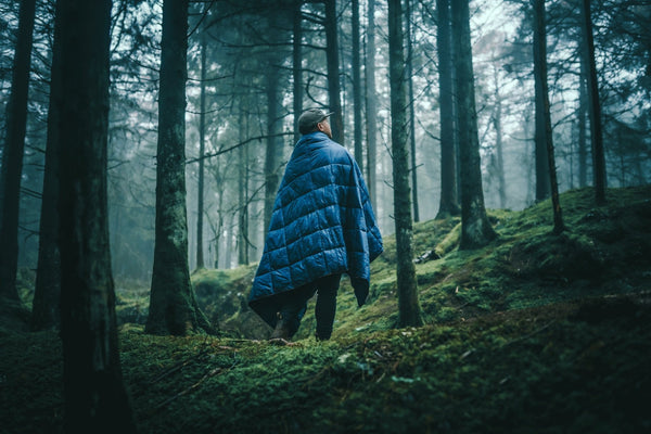 Image of a man wrapped in Nakie's Deep Ocean Blue Sustainable Down Puffy Blanket while standing in a campsite