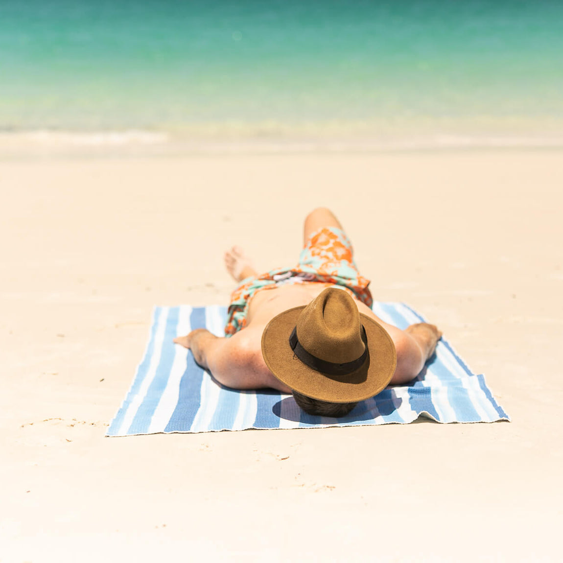 Blue and white striped recycled beach towel with person lying on sand wearing orange patterned swimwear and tan hat